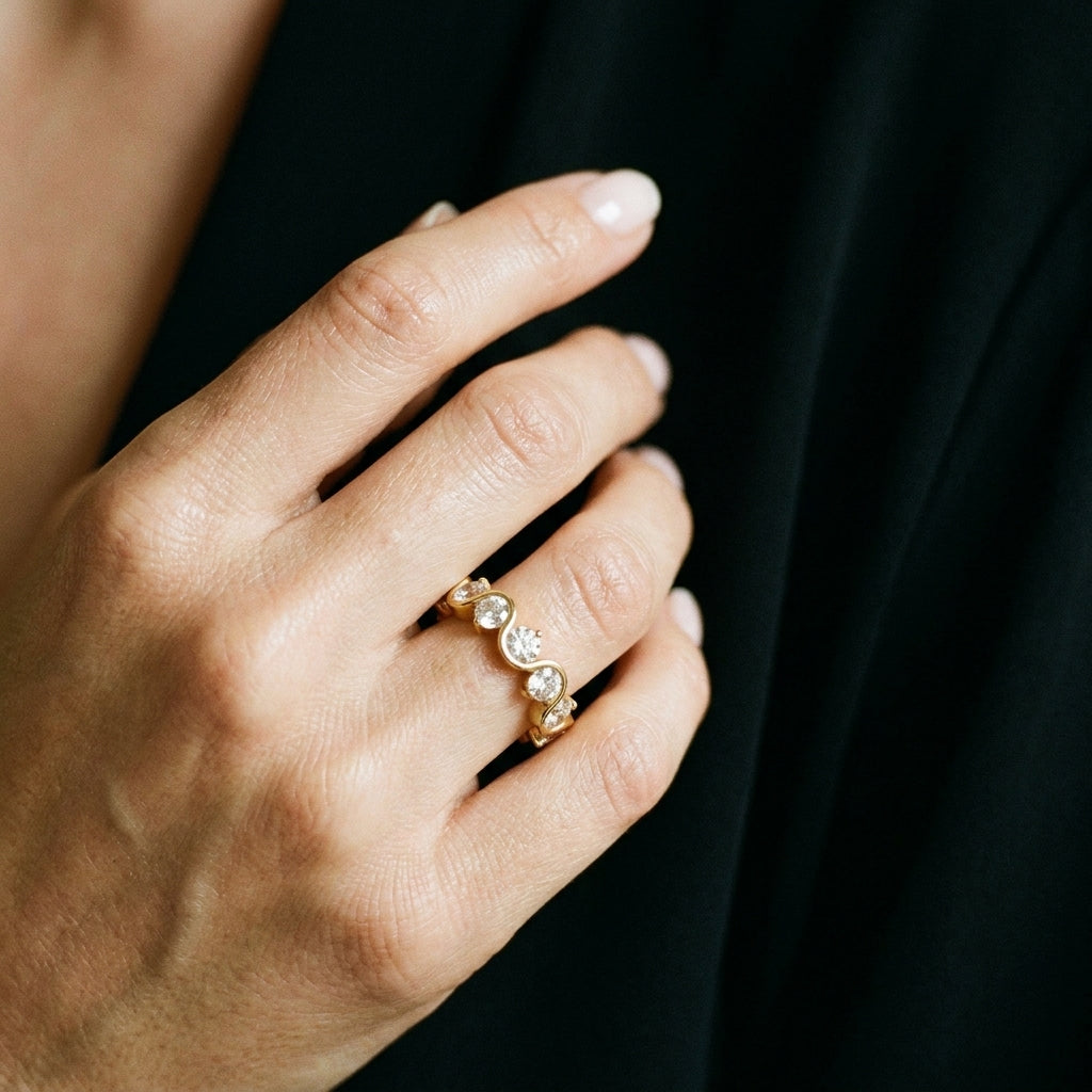 A close-up of a hand wearing a delicate gold ring with three small clear round gemstones, set against a dark black fabric background.