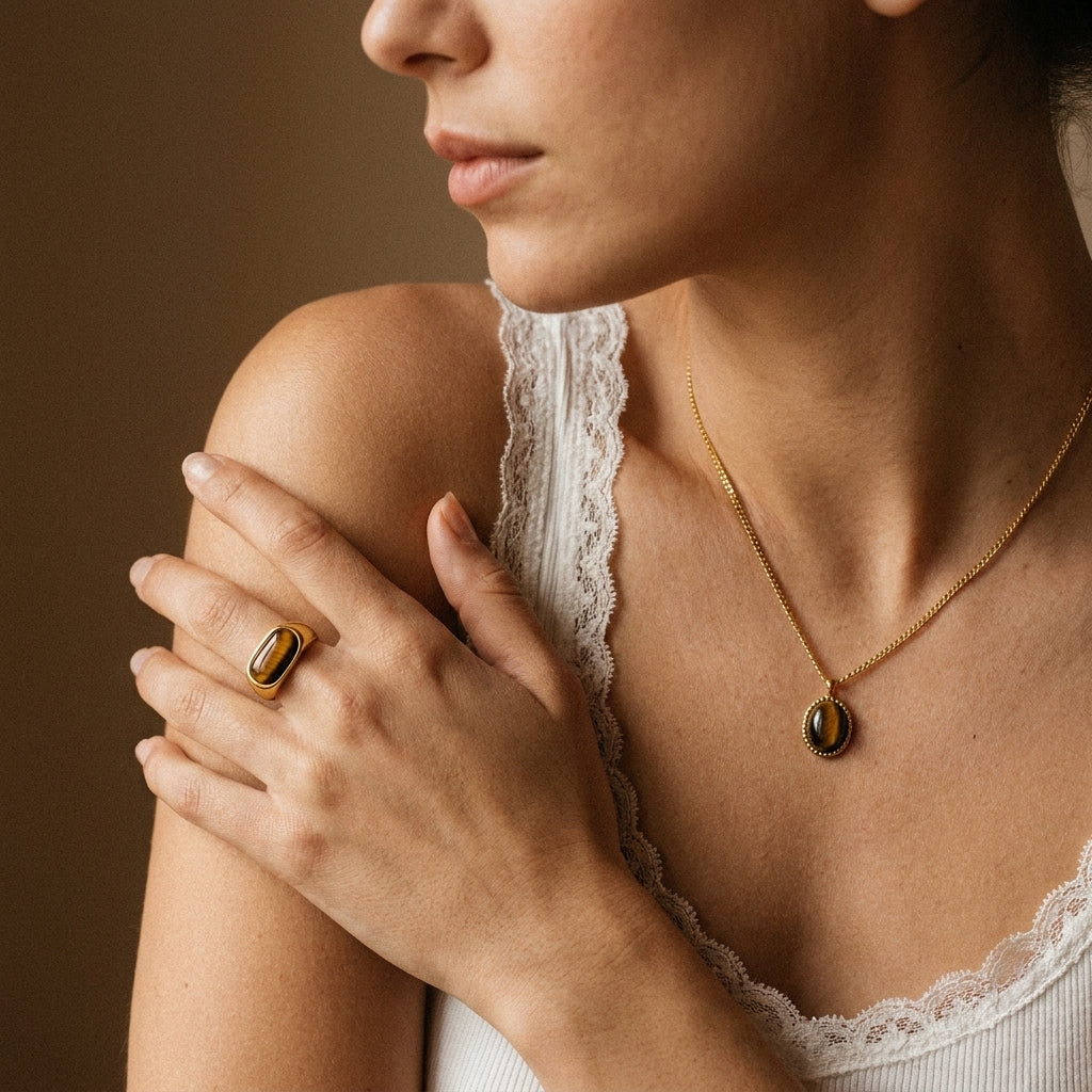 A close-up lifestyle shot of a woman wearing a gold ring with a brown oval stone on her finger and a matching gold necklace with a brown oval pendant. She is wearing a white lace-trimmed top against a warm brown background.