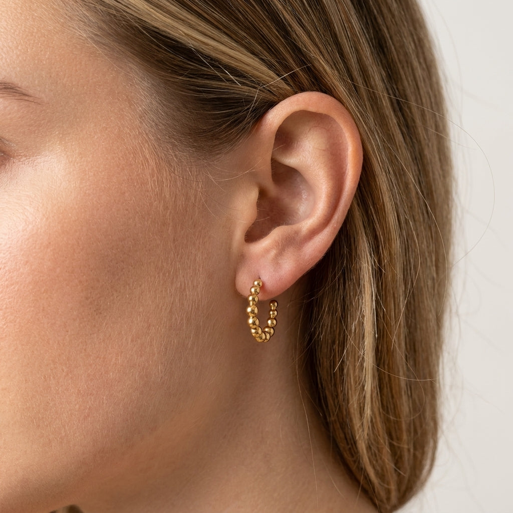 A close-up of a model's ear wearing a small gold huggie hoop earring with a textured beaded finish, set against a clean light gray background.