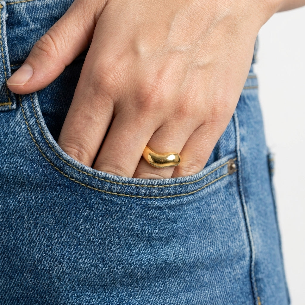 A close-up of a person's hand tucked into a blue denim pocket, wearing a smooth, polished gold ring.