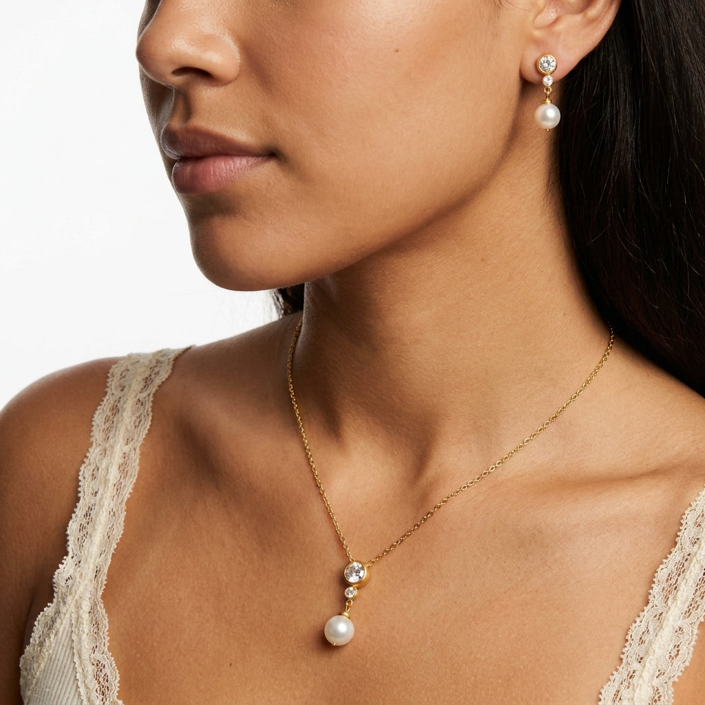 A close-up of a woman wearing a delicate gold necklace with a white pearl pendant and matching drop earrings with clear stones.