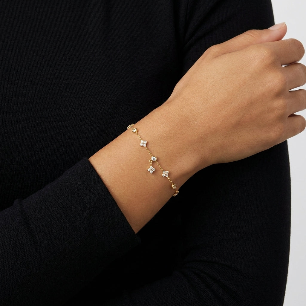 A delicate gold chain bracelet with small floral charms and clear stones, shown worn on a person's wrist against a black long-sleeve top.