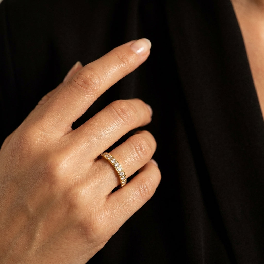 A close-up of a person's hand wearing a thin gold band ring set with small clear stones, against a dark black background.