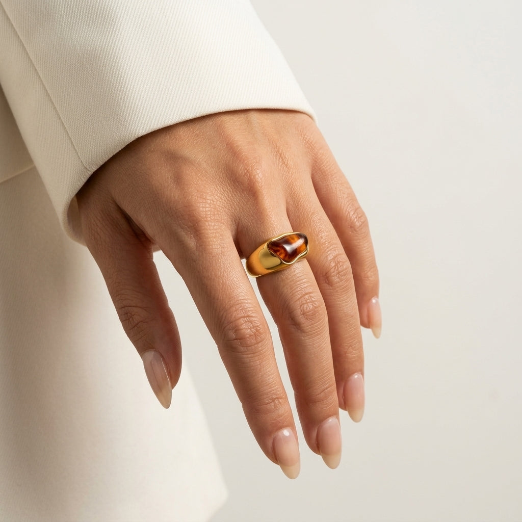 A close-up of a hand wearing a polished gold ring with a smooth, polished oval stone set against a soft, light cream background.