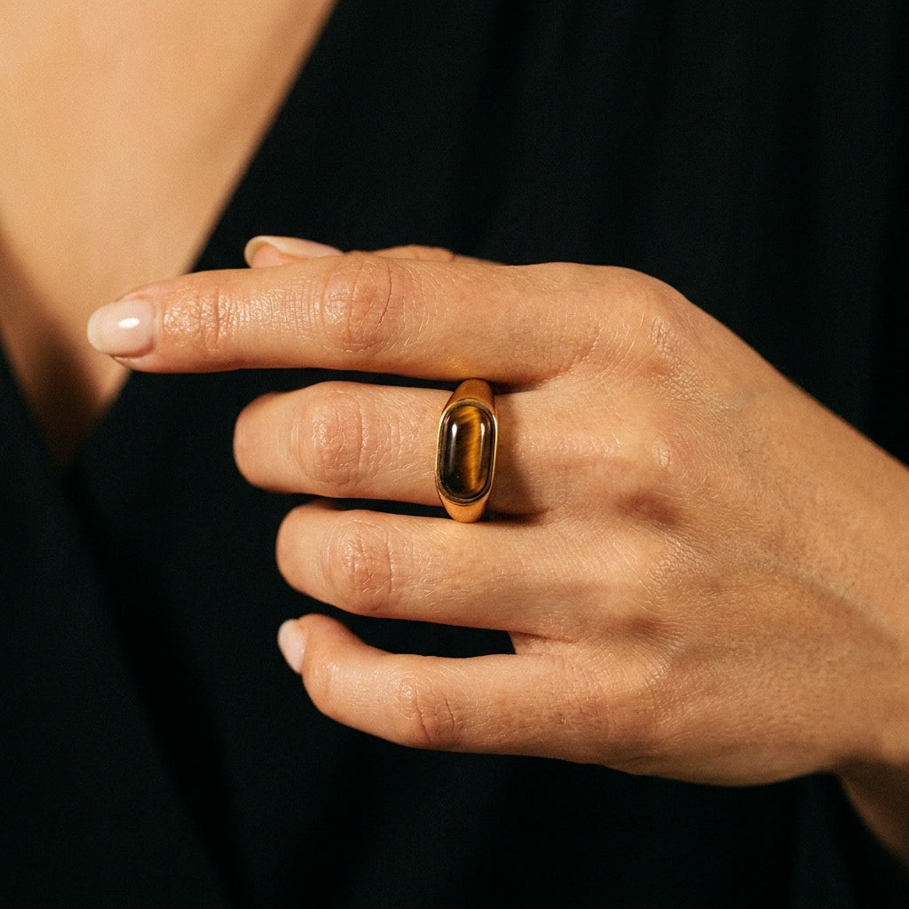 A close-up of a hand wearing a gold ring with a polished tiger's eye stone, set against a dark black top background.