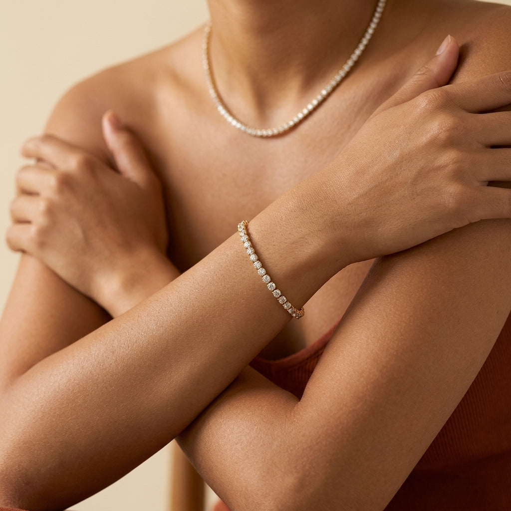 A close-up lifestyle shot of a model wearing a delicate gold bracelet adorned with small clear stones, paired with a matching pearl necklace, against a warm beige background.