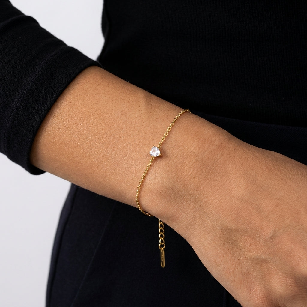 A delicate gold chain bracelet with a single clear crystal charm, shown worn on a person's wrist against a black top and light gray background.