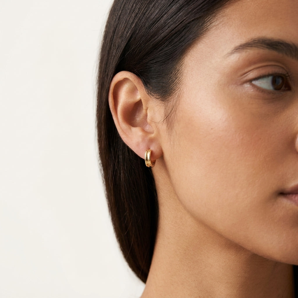A close-up of a woman's ear wearing a small gold hoop earring against a clean off-white background.