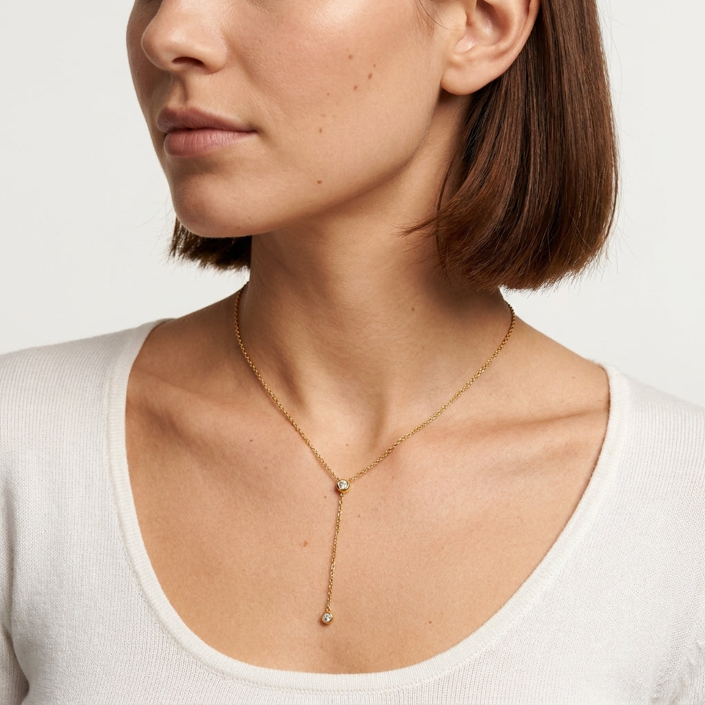 A close-up of a woman wearing a delicate gold Y-necklace with a small clear gemstone at the center and a longer drop ending in another small stone, paired with a white ribbed top against a light gray background.