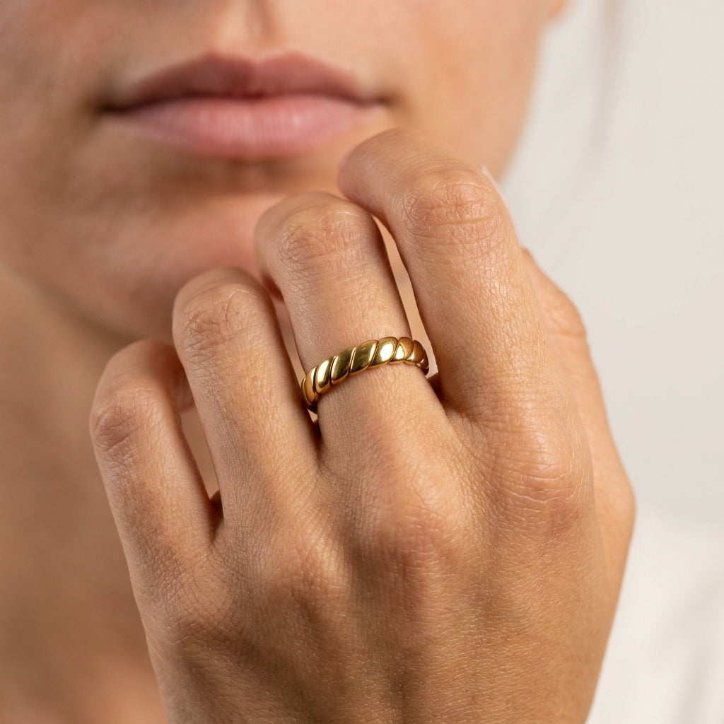 A close-up of a person's hand wearing a thin, twisted gold band ring, with their lips and lower face visible in the background.