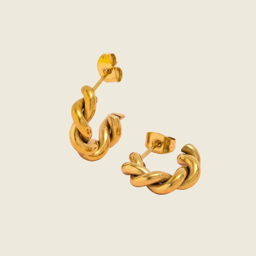A pair of gold huggie hoop earrings with a twisted, braided design, displayed on a soft pink background.