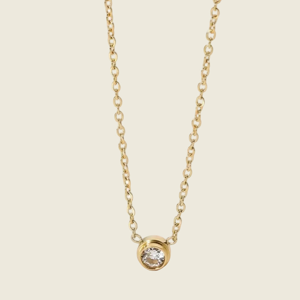 A delicate gold chain necklace with a small bezel-set clear gemstone pendant, displayed against a soft, pale pink background.