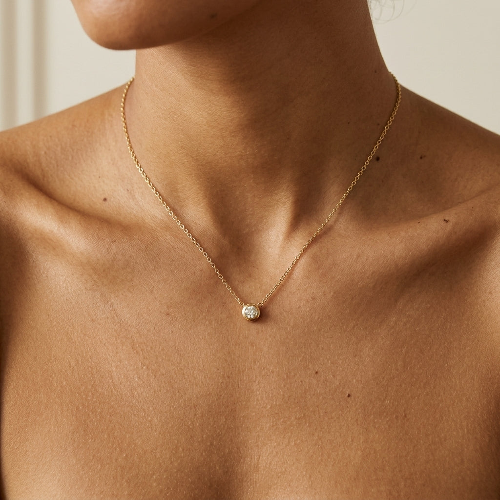 A close-up of a model's neck and upper chest wearing a delicate gold chain necklace with a small round clear gemstone pendant.