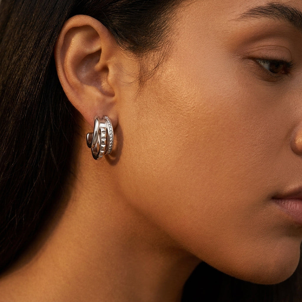 A close-up side profile of a woman wearing a silver hoop earring with textured, layered bands and small clear stones, set against a dark background.