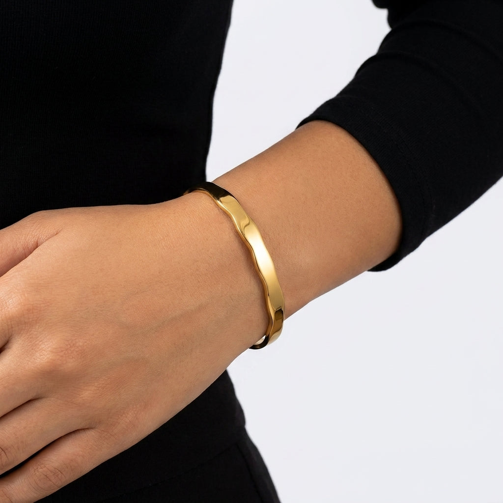 A polished gold metal bangle bracelet worn on a person's wrist, shown against a black long-sleeve top and a light gray background.