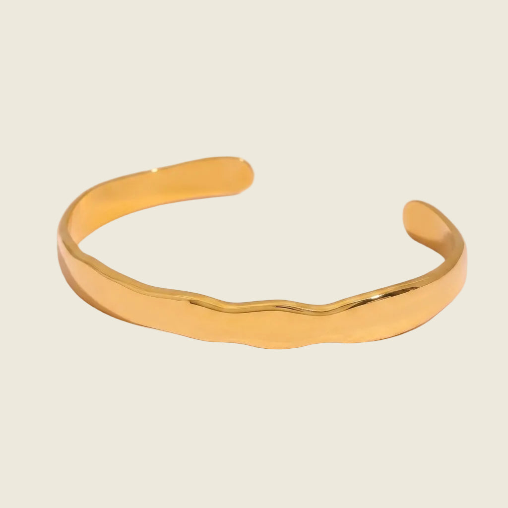 A polished gold cuff bracelet with a wavy, organic edge design, displayed on a soft pink background.