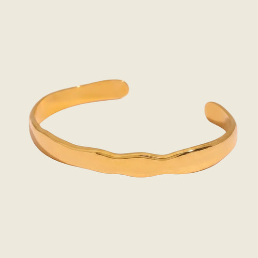 A polished gold cuff bracelet with a wavy, organic edge design, displayed on a soft pink background.