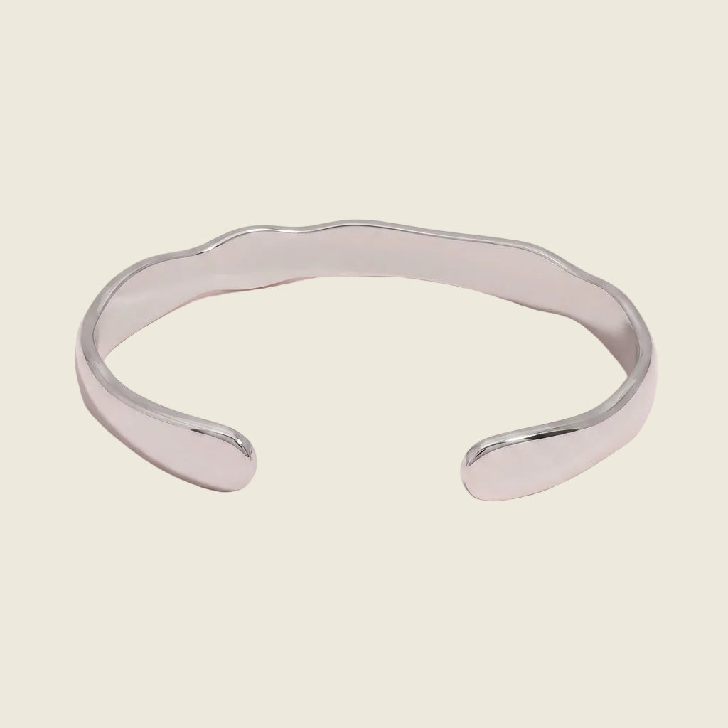 A polished silver cuff bracelet with a wavy, organic edge, displayed on a soft pink background.