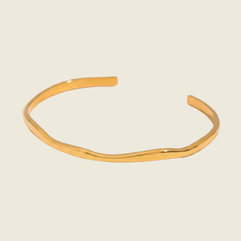 A polished gold wavy cuff bracelet on a light pink background.