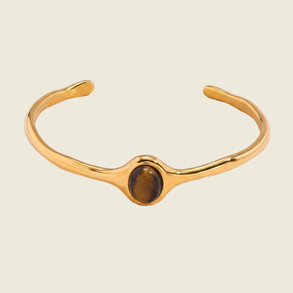 A polished gold-tone cuff bracelet featuring a central bezel-set oval tiger's eye stone, displayed on a soft pink background.