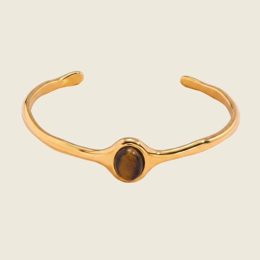 A polished gold-tone cuff bracelet featuring a central bezel-set oval tiger's eye stone, displayed on a soft pink background.