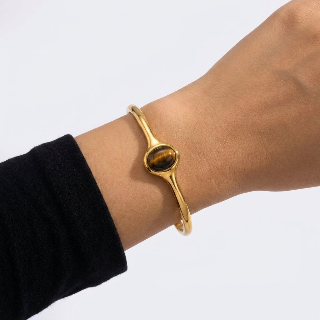 A gold-toned bangle bracelet with a round tiger's eye stone, shown worn on a person's wrist against a plain light background.
