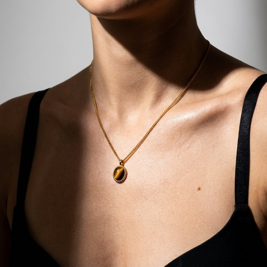 A close-up of a model wearing a delicate gold chain necklace with an oval pendant featuring a brown and gold tiger's eye pattern, styled with a black spaghetti strap top.