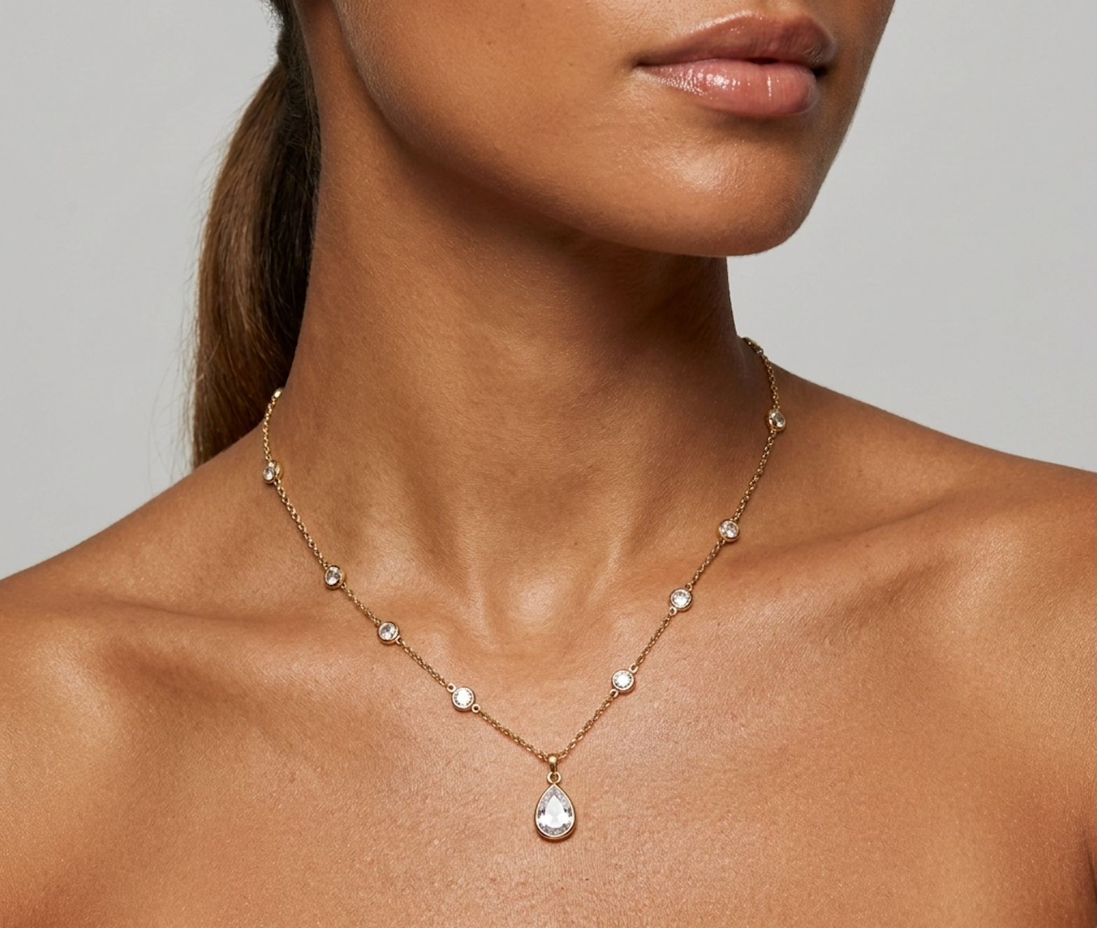 A close-up of a model wearing a delicate gold chain necklace with small clear stones and a teardrop-shaped clear pendant, shown against a neutral gray background.
