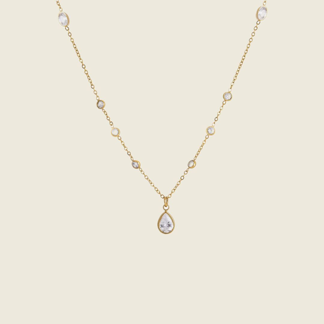 A delicate gold chain necklace featuring a teardrop-shaped clear gemstone pendant and small round clear gemstones spaced along the chain, set against a solid light beige background.