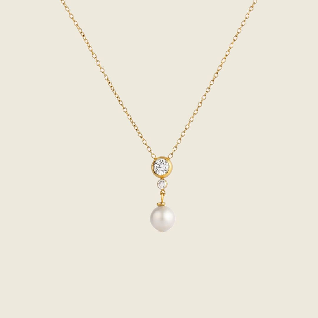 A delicate gold chain necklace featuring a pendant with a small round clear stone, a tiny clear accent stone, and a dangling white pearl, set against a solid light beige background.