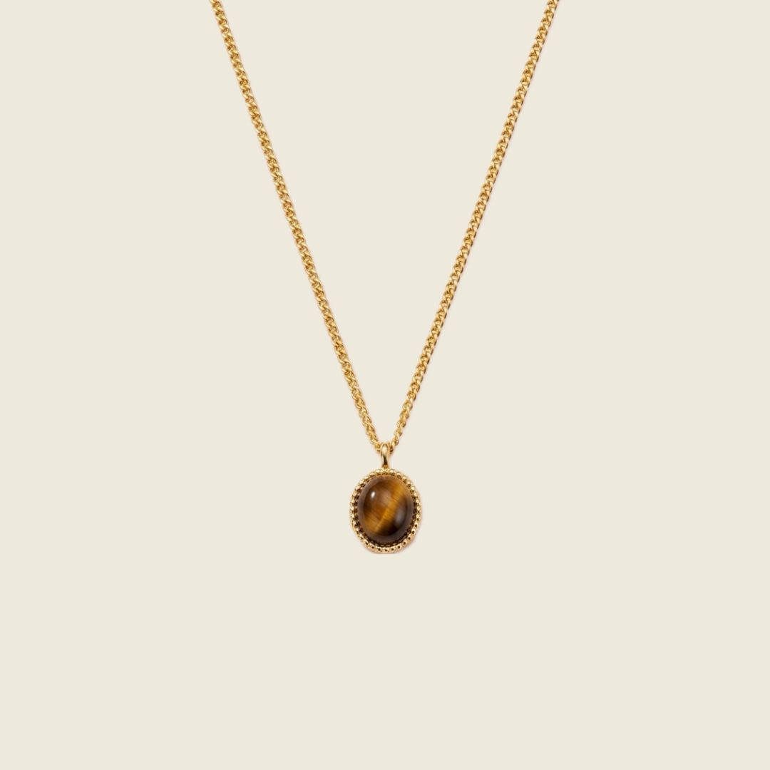 A gold chain necklace with an oval pendant featuring a brown and gold tiger's eye stone, set against a solid light beige background.