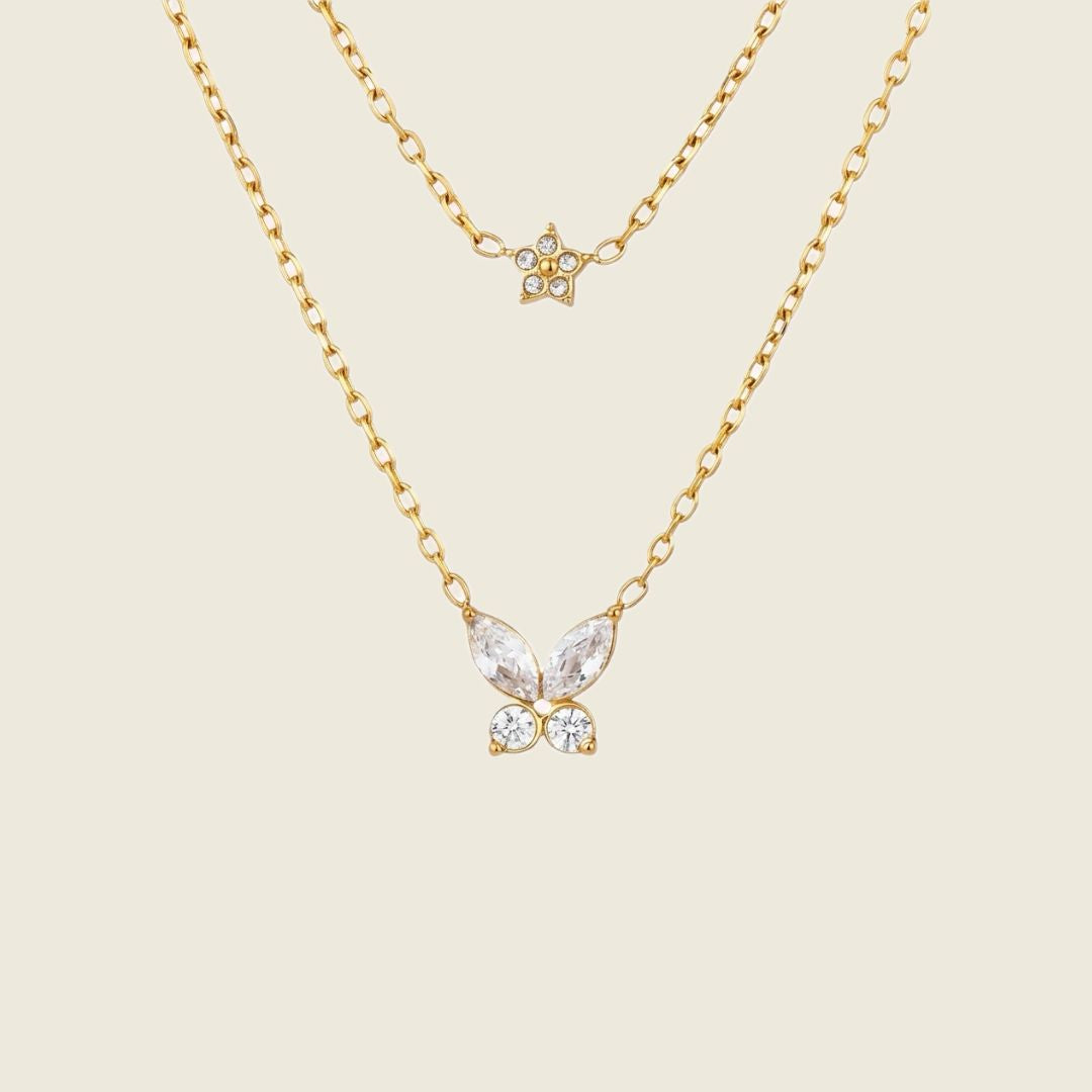 Two layered gold necklaces on a light beige background. The shorter necklace features a small gold star with clear rhinestones, while the longer necklace has a butterfly pendant with marquise-cut clear crystals and round clear crystals.