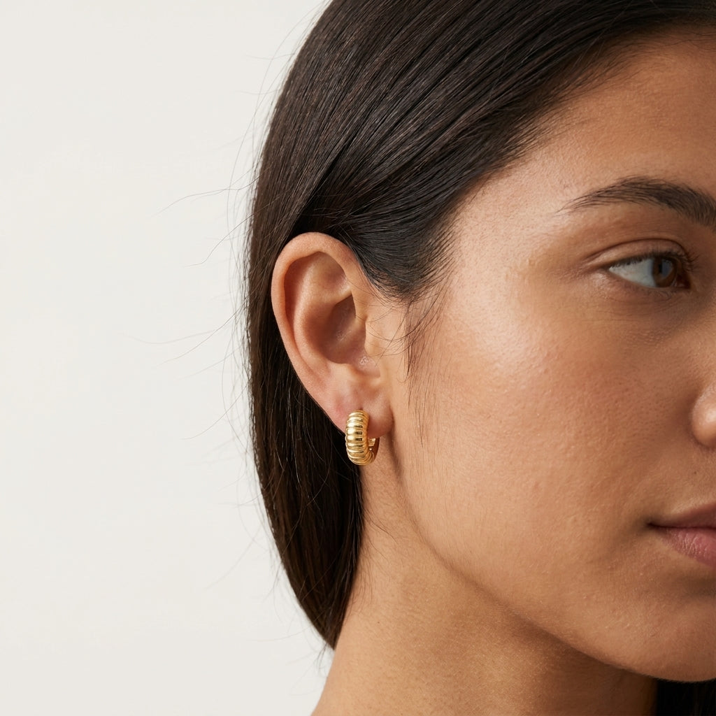 A close-up side profile of a woman wearing a small gold hoop earring with a textured, ridged outer edge, set against a plain light background.